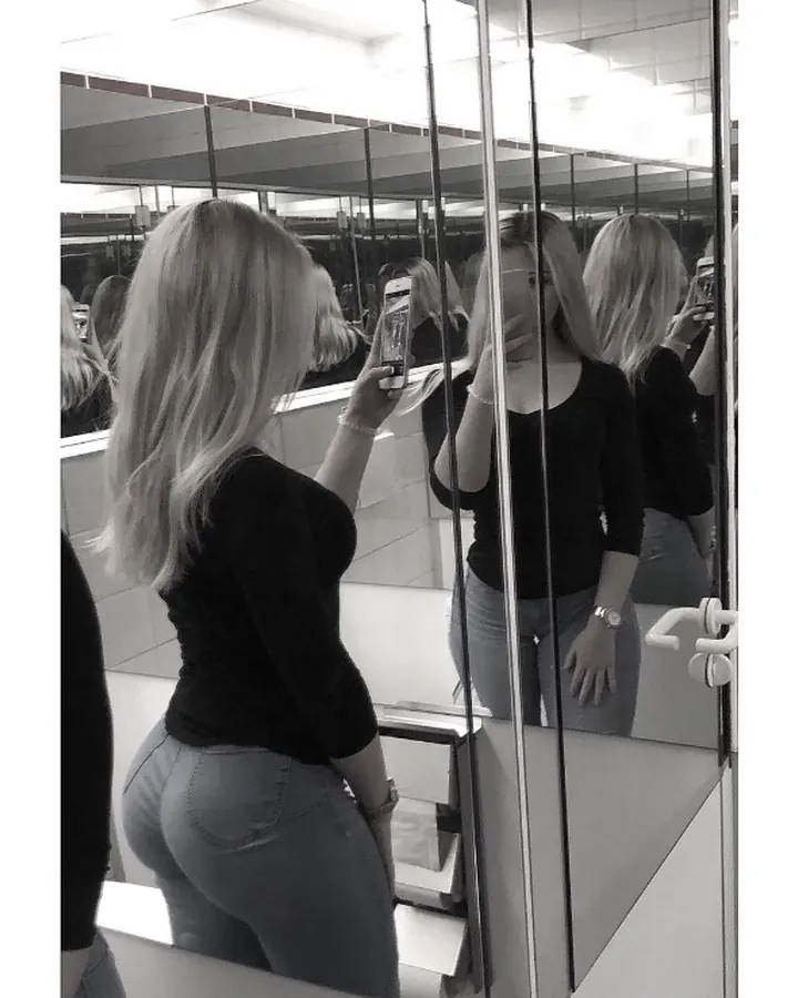 Femme aux cheveux blonds platine assise sur un canapé, vêtue d'une robe noire, avec des chaussures à talons.