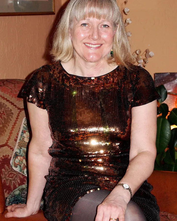 Femme aux cheveux blonds platine assise sur un canapé, vêtue d'une robe noire scintillante, avec des collants noirs et des chaussures noires.