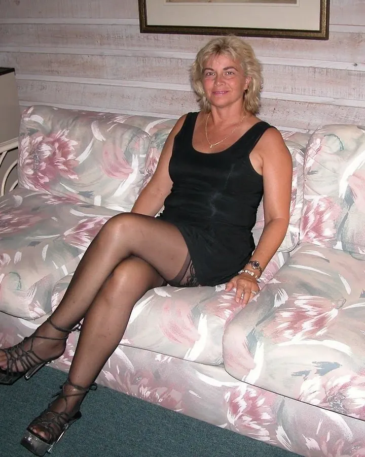 Femme aux cheveux blonds platine assise sur un canapé à motifs floraux, vêtue d'une robe noire courte et de bas noirs, avec des chaussures à talons.
