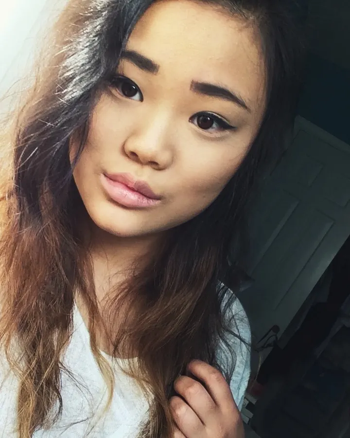 Femme asiatique aux cheveux châtains en chemise blanche, prenant un selfie dans une chambre, regardant l'appareil.