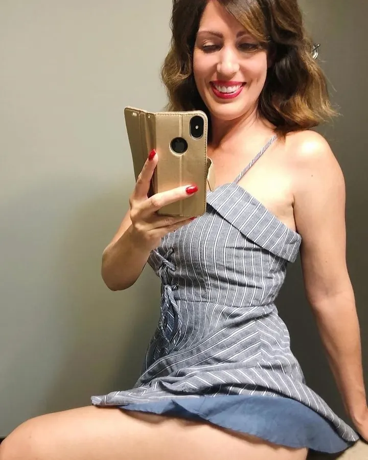 Femme aux cheveux châtains en robe rayée grise et bleue, assise sur un canapé, prenant un selfie avec un téléphone doré.