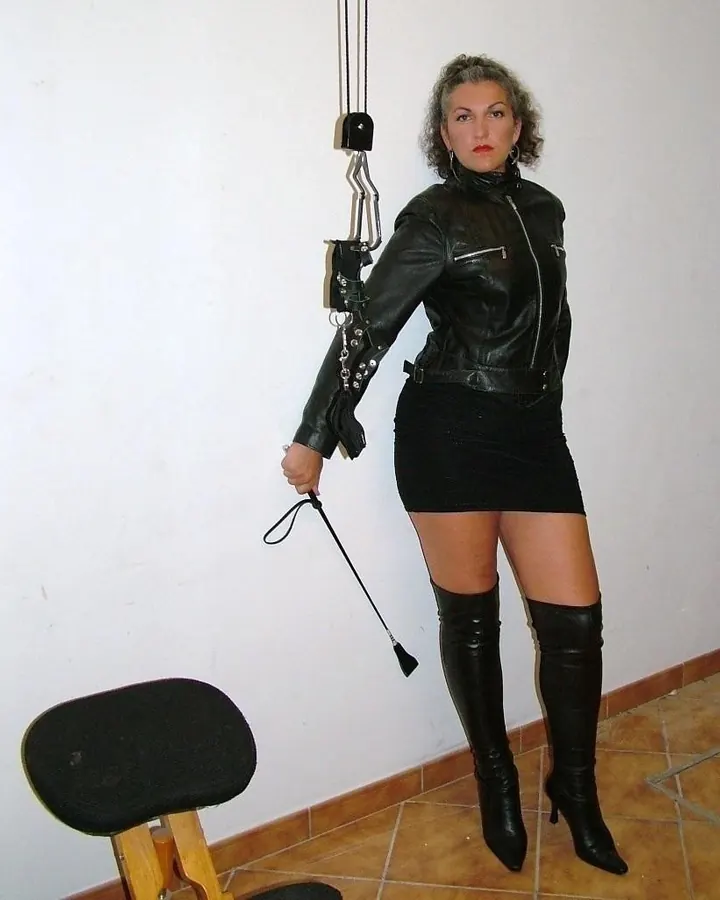 Femme aux cheveux gris clair en veste en cuir noir, jupe courte noire et bottes hautes, tenant un fouet, devant un mur blanc.