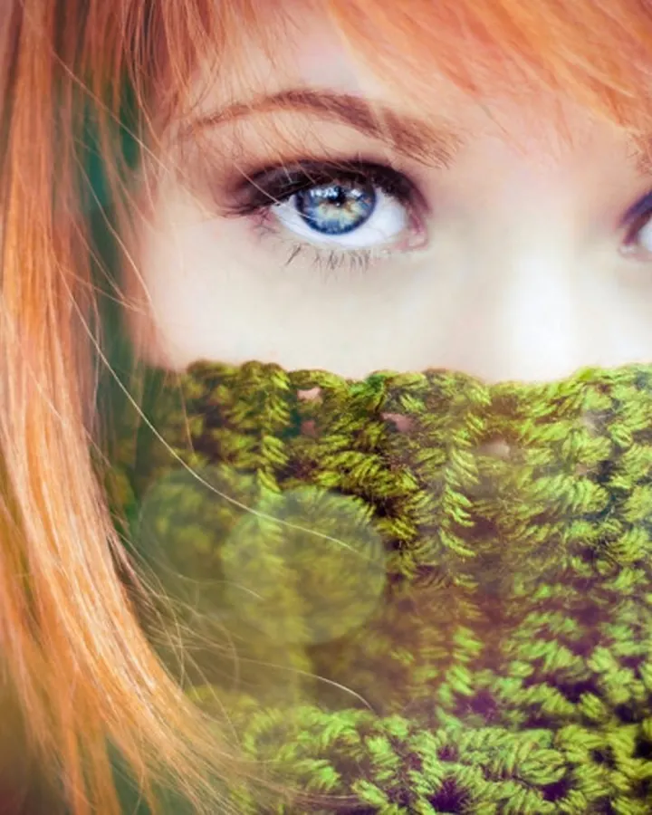 Jeune femme rousse aux yeux bleus, pull vert tricoté couvrant le bas du visage, regard vers l'objectif
