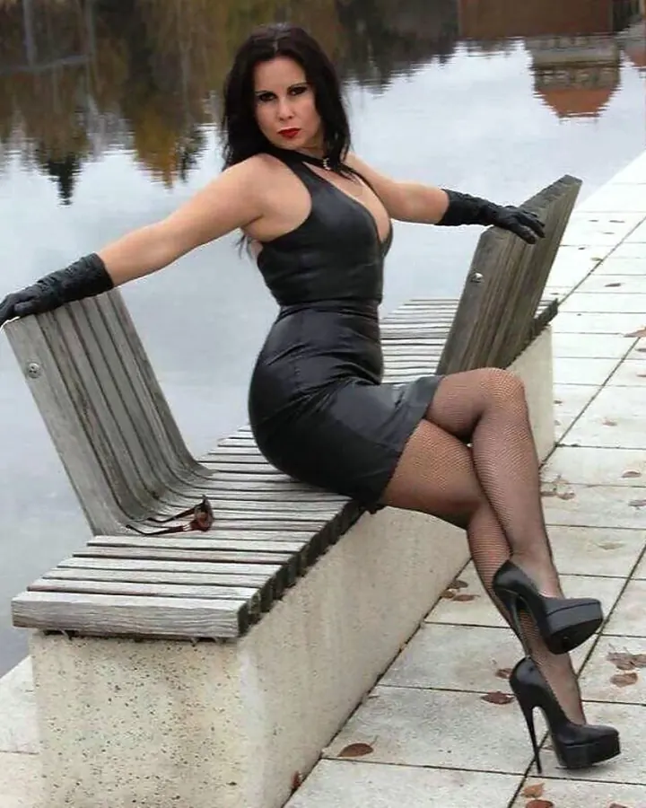 Femme brune en robe noire et gants, assise sur un banc près d'un plan d'eau