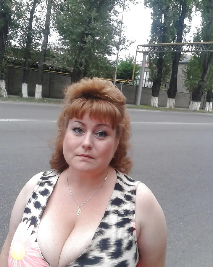 Femme en robe à imprimé léopard rouge, coiffure blonde bouclée, assise sur un banc en bois, tenant une cigarette.