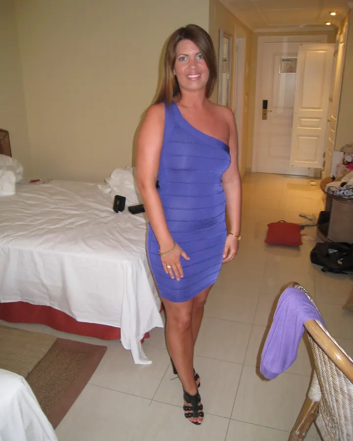 Femme portant une robe bleue asymétrique, debout dans une chambre avec un lit et des bagages en arrière-plan.