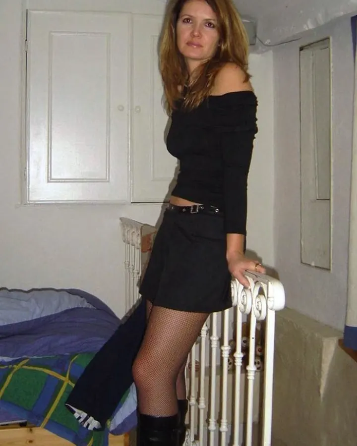 Femme portant une tenue noire décolletée, posant dans une chambre avec un lit et un radiateur en arrière-plan.