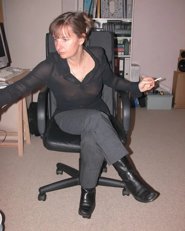 Femme européenne assise dans un fauteuil noir, en chemise noire transparente, tenant une cigarette, dans un bureau.