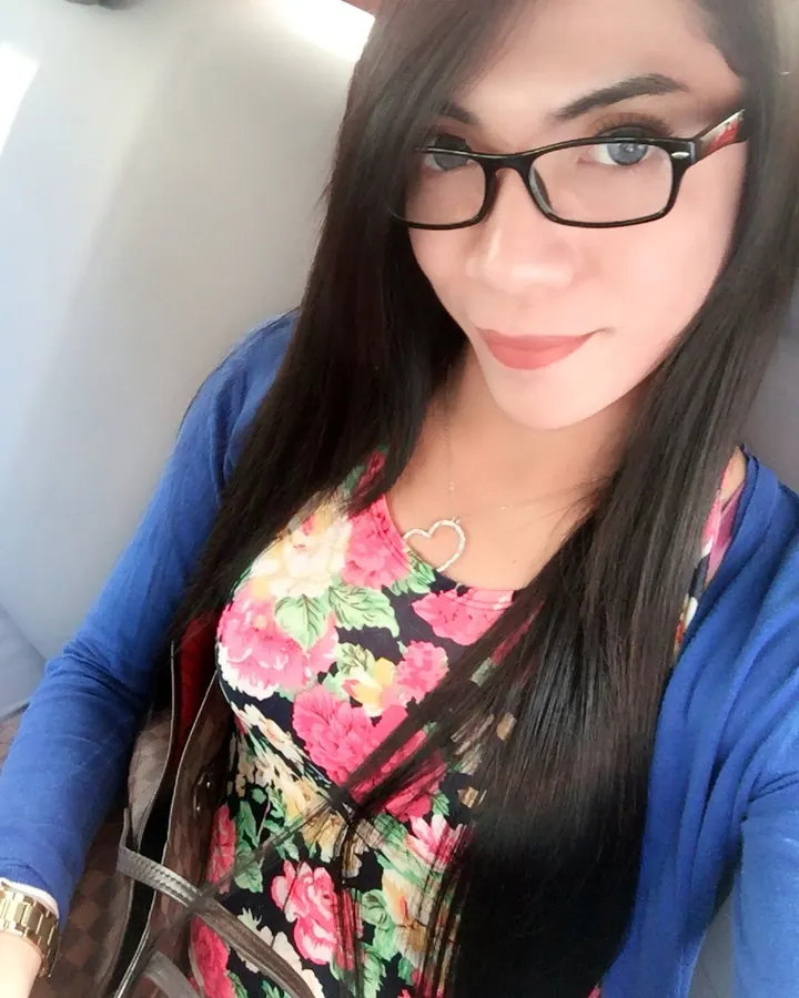 Femme latino portant des lunettes, en chemisier floral et cardigan bleu, assise dans une voiture, souriant légèrement.