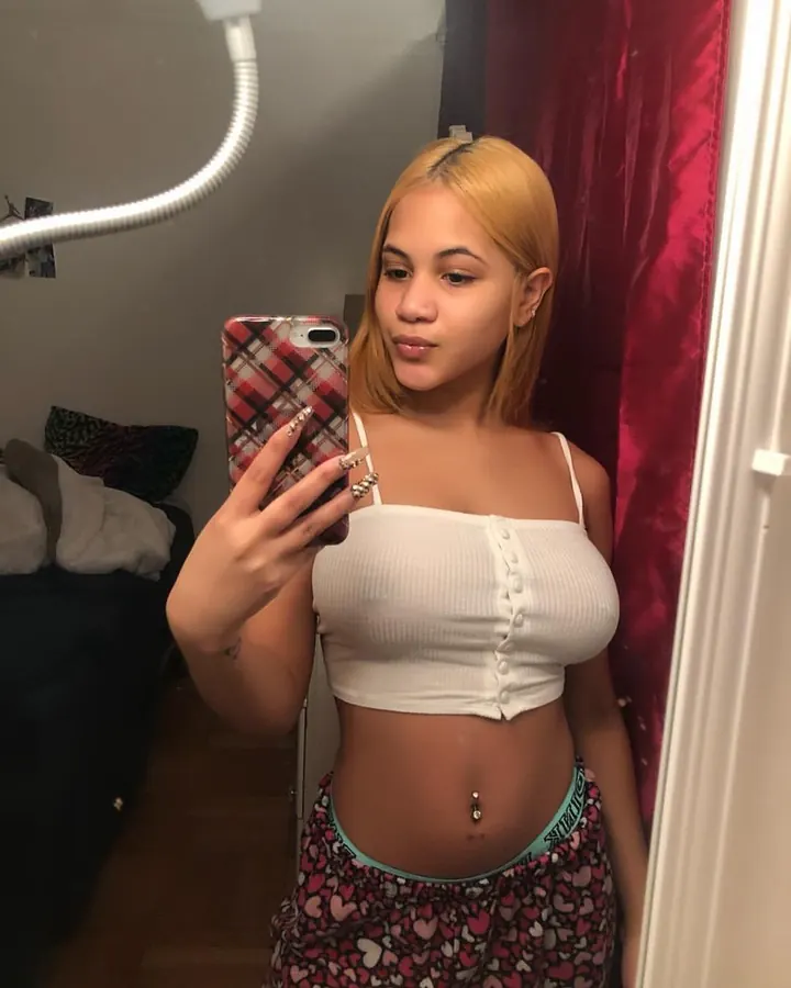 Femme aux cheveux foncés, portant un haut blanc et un short noir, prenant un selfie devant un miroir, dans une chambre.
