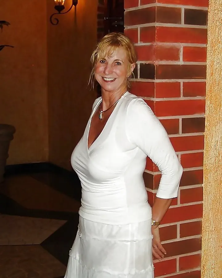 Femme blonde, assise sur un canapé, portant une robe blanche, souriante, avec un mur de briques en arrière-plan.