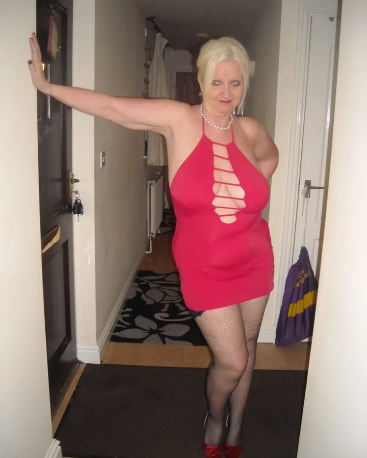 Femme aux cheveux blonds, portant une robe rouge, debout dans un couloir, souriante, avec un sac à main violet.