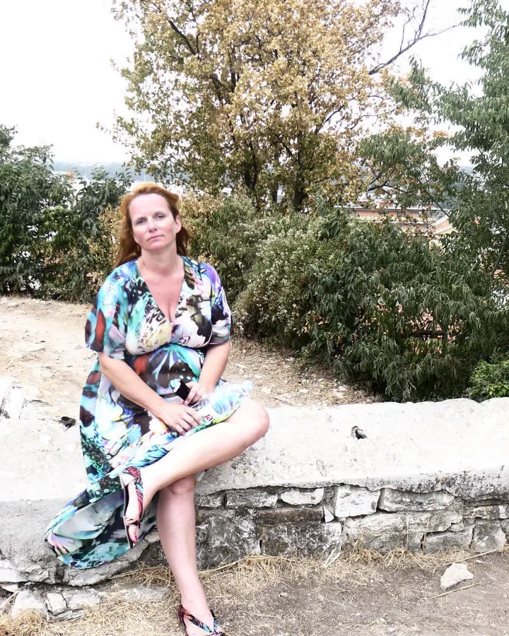 Femme aux cheveux roux, vêtue d'une robe colorée, assise sur un mur de pierre, regardant l'appareil photo, avec des arbres en arrière-plan.