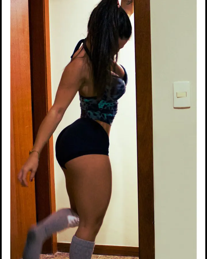 Femme aux cheveux bruns, portant un haut blanc et un short noir, prenant un selfie devant un miroir, dans une chambre.