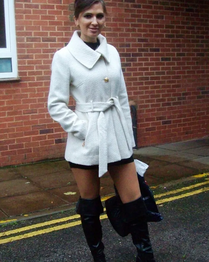 Femme souriante en manteau blanc, jupe courte noire et bottes noires, debout devant un mur de briques