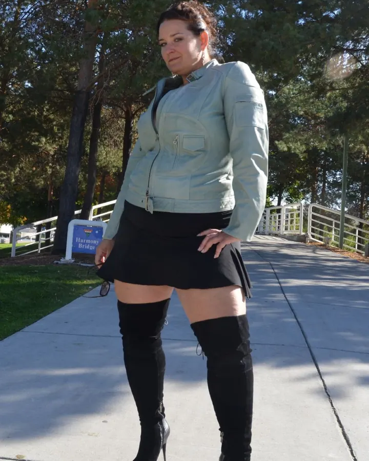 Femme en veste claire, jupe noire et bottes hautes, debout sur un trottoir ensoleillé