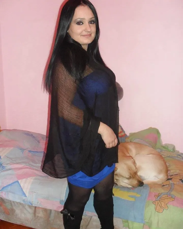 Femme aux cheveux bruns-noirs portant une robe bleue sous une étole noire transparente, debout près d'un lit où dort un chien.
