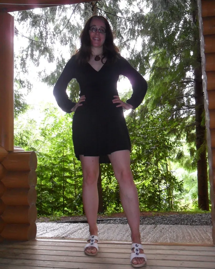 Femme aux cheveux châtains portant une robe noire, debout sur une terrasse en bois devant un arrière-plan verdoyant.