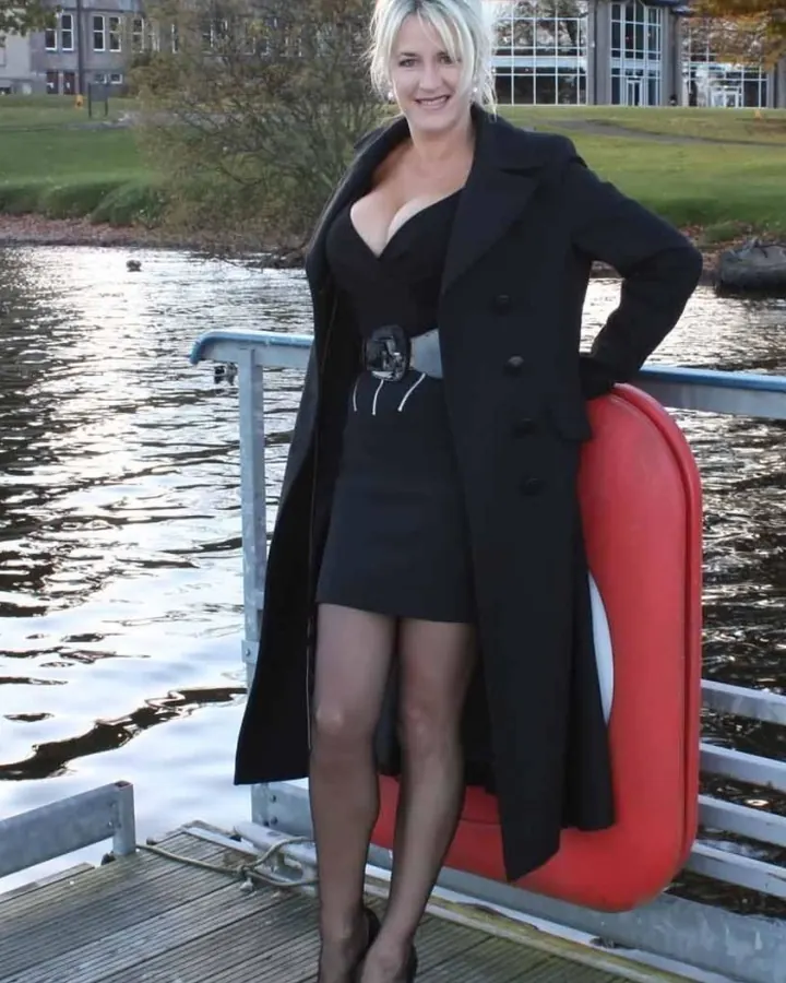 Femme aux cheveux gris platine portant un haut noir et un pantalon en cuir noir, assise sur un canapé bleu avec des bottes noires à talons.