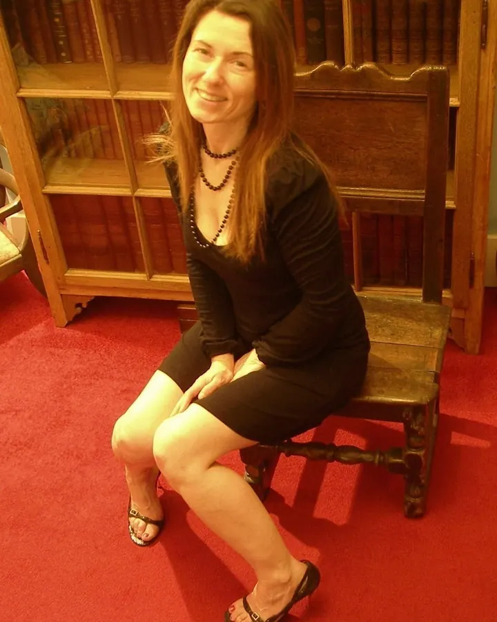 Femme aux cheveux châtains portant une robe noire en velours avec des accents roses, assise sur un canapé avec des bas noirs et des chaussures rouges brillantes.
