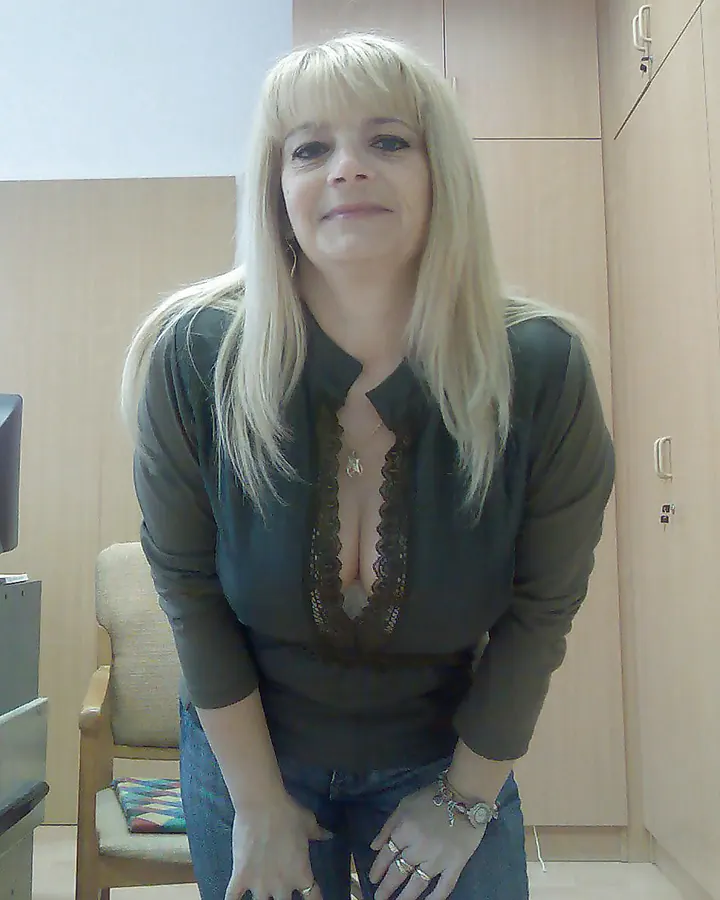 Femme aux cheveux gris platine portant un haut noir et un pantalon en cuir noir, assise sur un canapé bleu avec des bottes noires à talons.