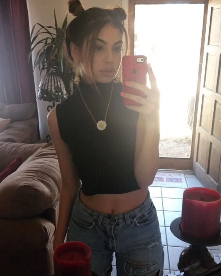 Jeune femme aux cheveux châtains coiffés en deux chignons, portant un haut noir et un jean déchiré, prenant un selfie dans un miroir avec des bougies rouges autour d’elle.