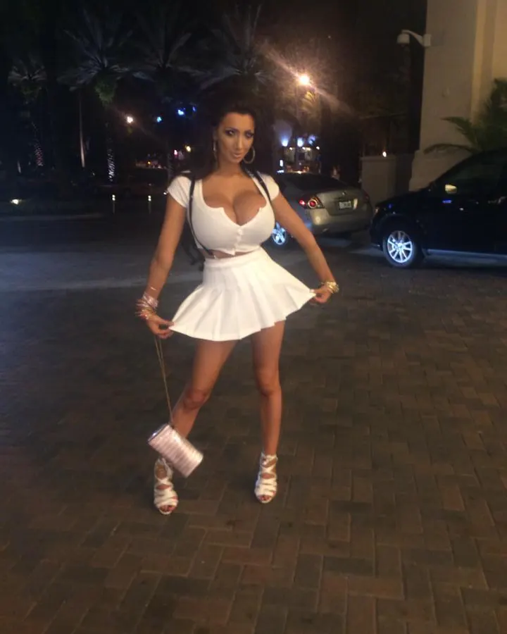 Femme aux cheveux noirs, portant un haut blanc et une jupe blanche courte, debout sur un trottoir pavé la nuit, tenant un sac à main blanc.