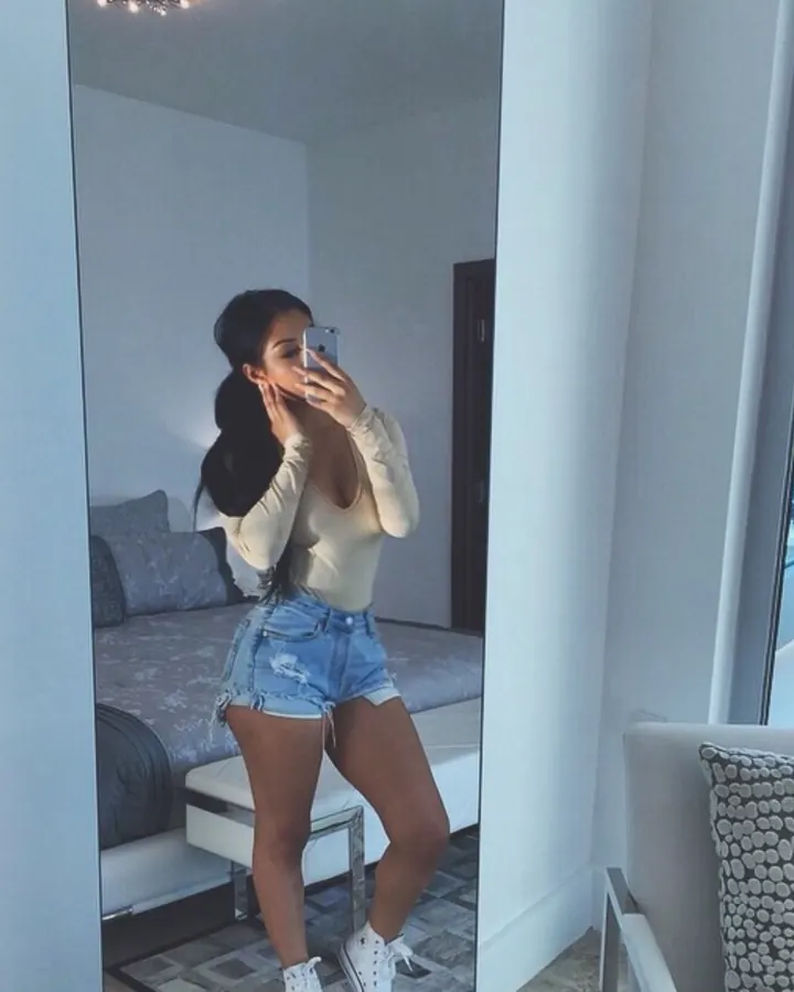 Femme aux cheveux noirs, portant un haut noir transparent et un haut beige, prenant un selfie devant un miroir dans une chambre moderne.