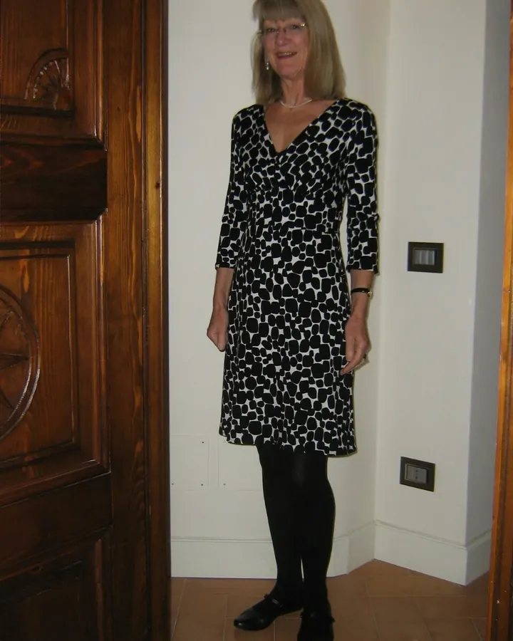 Femme blonde, portant un haut blanc, debout près d'un escalier en bois, souriant à l'appareil photo.
