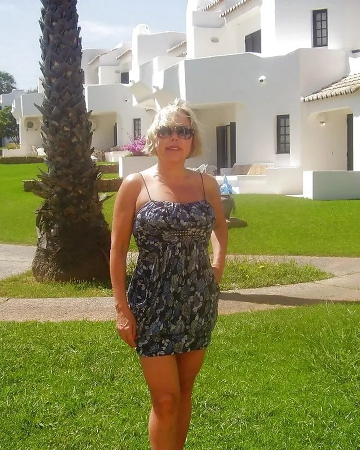 Femme blonde, portant une robe bleue à motifs, debout sur une pelouse verte devant des bâtiments blancs, sous un ciel clair.
