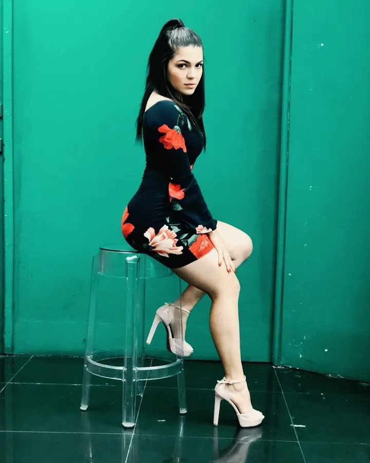 Femme aux cheveux noirs en queue-de-cheval, vêtue d'une robe noire à fleurs rouges, assise sur un tabouret transparent devant un mur vert