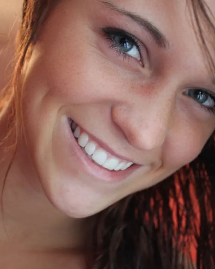 Femme souriante aux yeux bleus, avec des cheveux bruns, regardant directement l’appareil photo dans une lumière chaude.