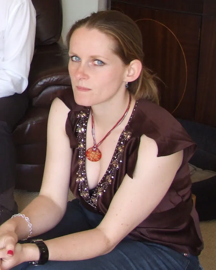 Femme aux yeux bleus, portant un haut marron orné de perles et un collier rouge, assise sur un canapé en cuir.