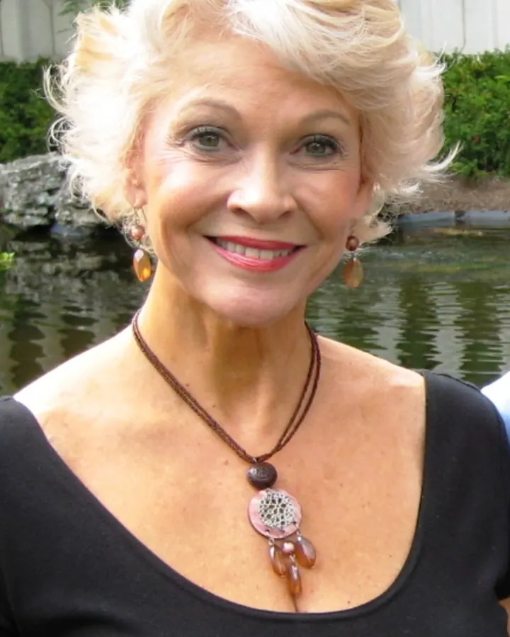 Femme aux cheveux blancs bouclés, portant un haut noir et un collier décoratif, souriant devant un étang.