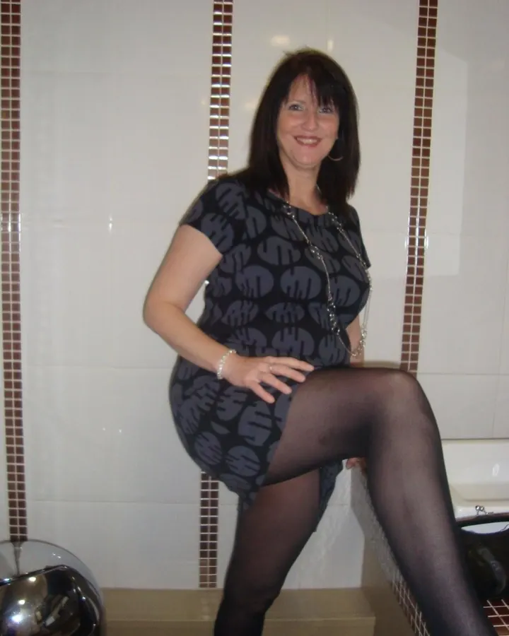 Femme en robe noire à motifs gris, portant des collants noirs, assise sur un rebord de baignoire dans une salle de bain.