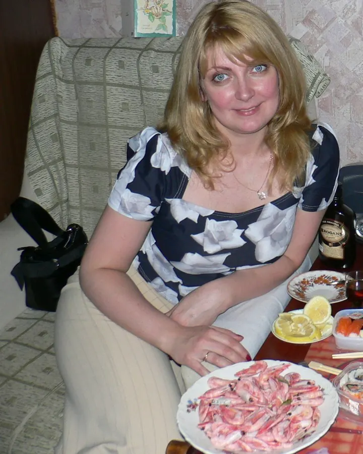 Femme blonde en haut blanc, assise à une table avec des crevettes, souriant, dans un cadre domestique