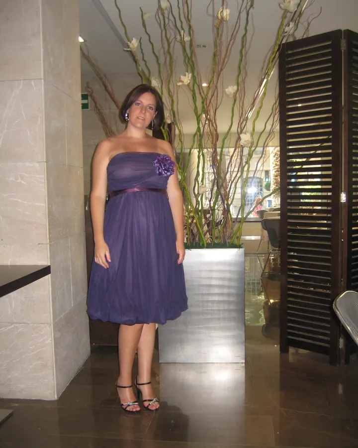 Femme en robe violette sans bretelles, fleur violette, cheveux bruns, souriant dans un hall