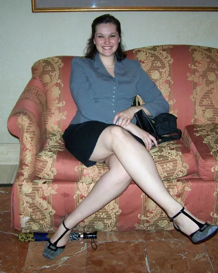 Femme aux cheveux châtains assise sur un canapé, chemise grise, jupe noire et chaussures à talons.
