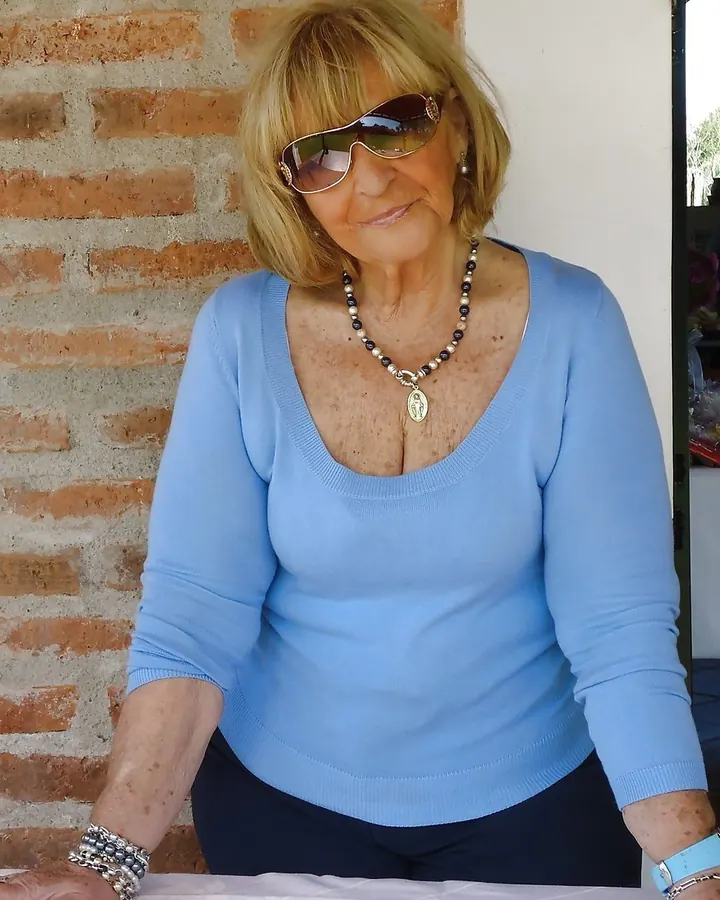 Femme blonde en pull bleu, assise près d'un mur en briques, lunettes de soleil.