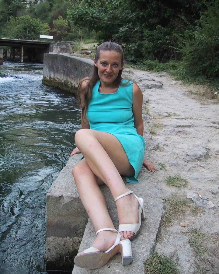 Femme aux cheveux châtains, vêtue d’une robe turquoise, assise sur un bord de rivière, souriant à la caméra.