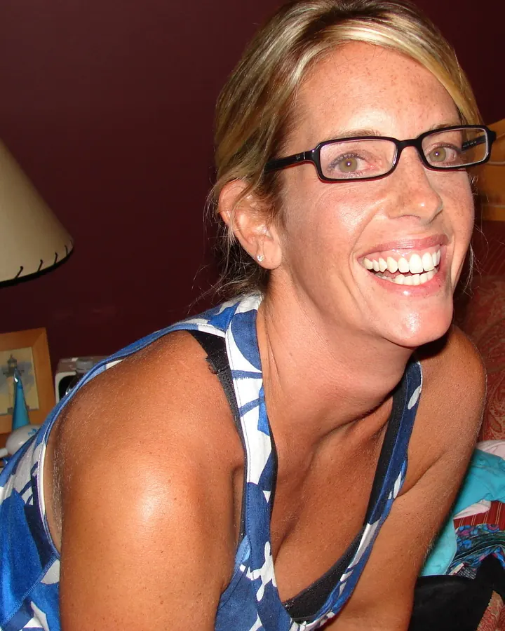 Femme aux cheveux blonds, portant des lunettes, en débardeur bleu et blanc, souriant largement