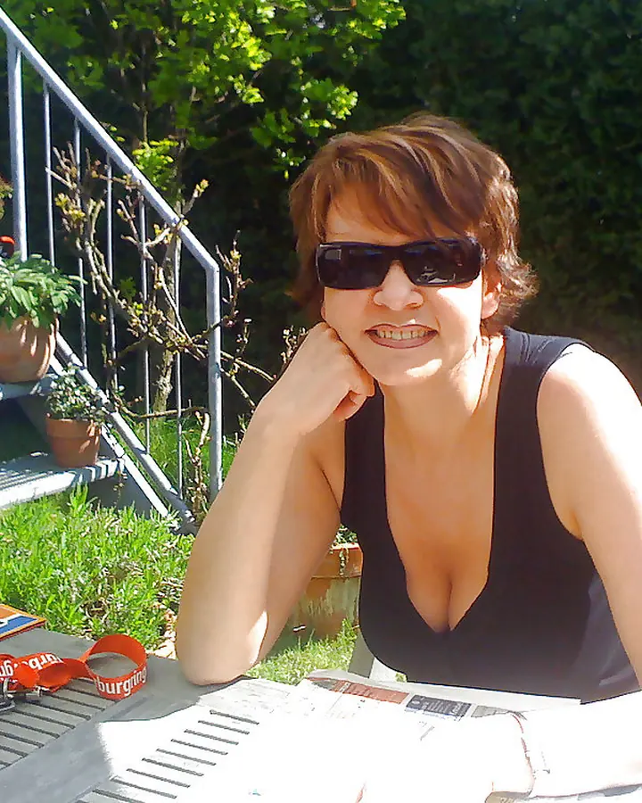 Femme aux cheveux châtains portant des lunettes de soleil, assise à une table en extérieur, vêtue d’un haut noir décolleté, entourée de verdure.
