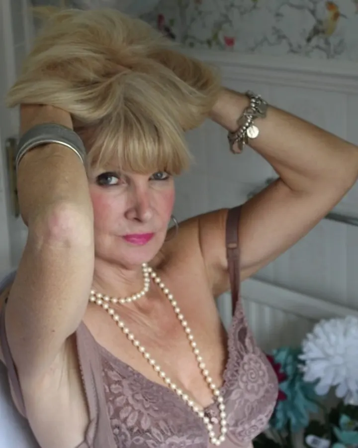 Femme blonde portant une robe rose, les mains dans les cheveux, assise sur une chaise dans une pièce lumineuse.