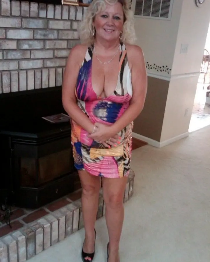 Femme blonde portant une robe colorée, souriante, debout devant une cheminée en briques.