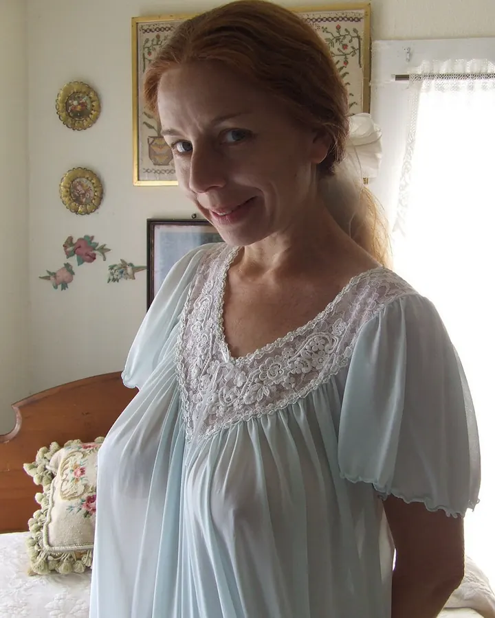 Femme aux cheveux roux portant une chemise de nuit bleu clair avec dentelle blanche, souriant légèrement dans une chambre décorée avec des motifs floraux.