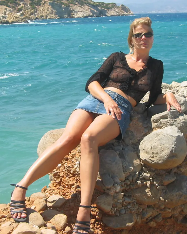 Femme blonde en haut noir transparent et short en jean, assise sur un mur de pierres près de la mer, portant des lunettes de soleil.