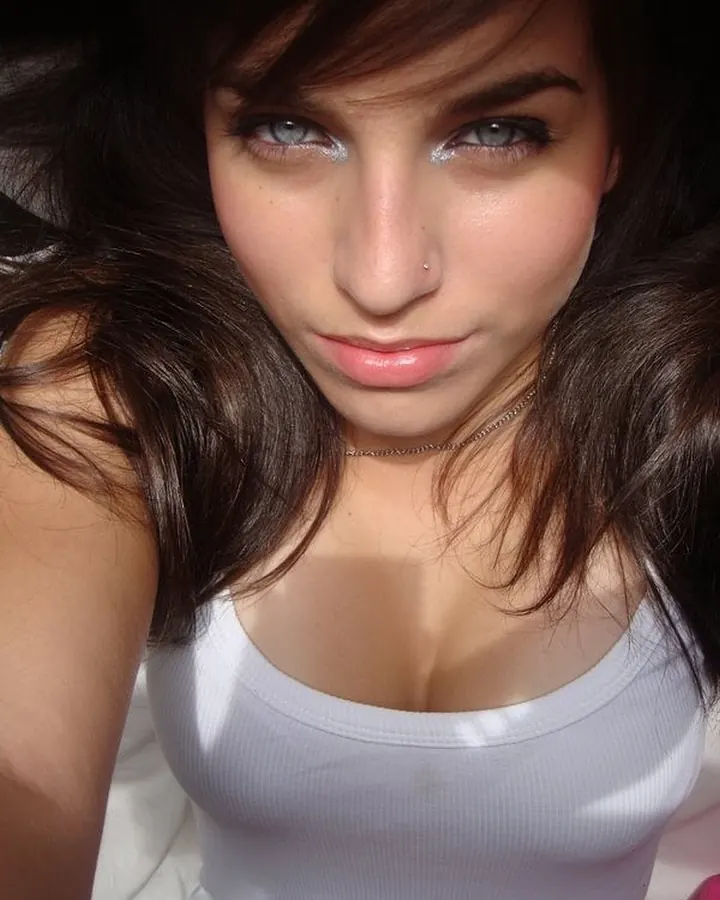 Femme brune en gros plan, regard vers l’appareil, débardeur blanc, maquillage lumineux.