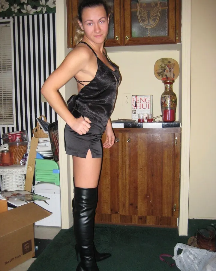 Femme blonde en top noir et short, bottes noires, pose debout.