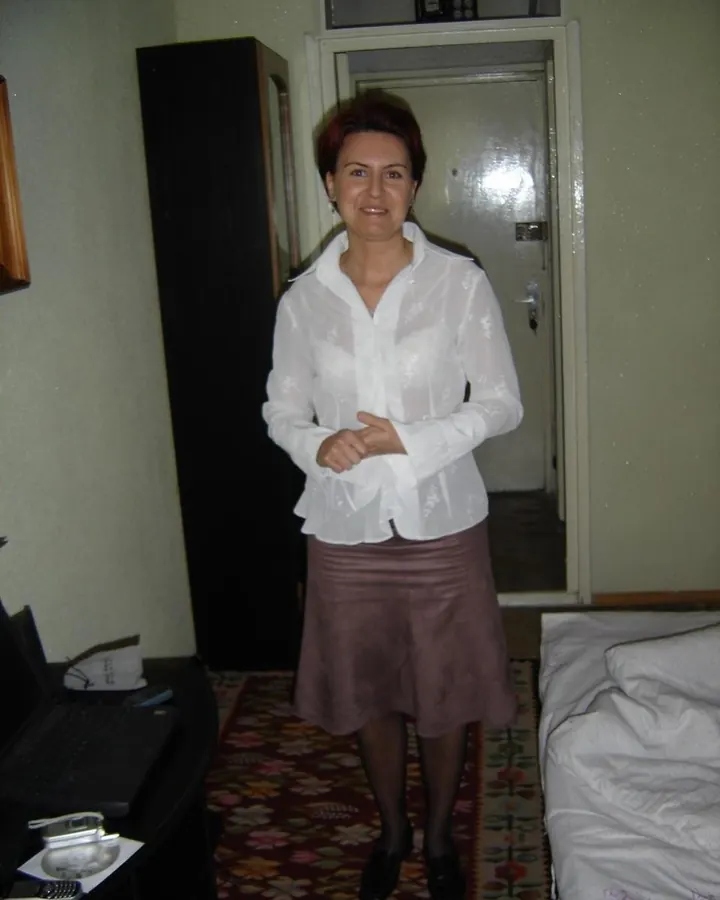 Femme aux cheveux courts roux, en chemise blanche transparente et jupe marron, debout dans une chambre.