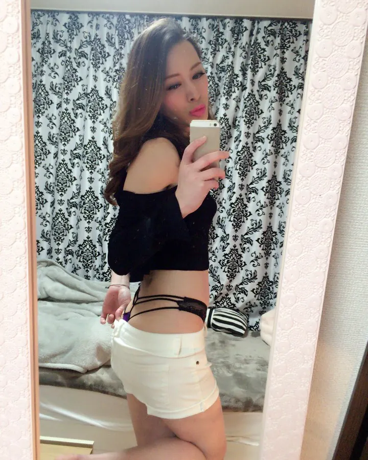 Femme asiatique aux cheveux bruns, en haut noir et short blanc, prenant un selfie devant un miroir.
